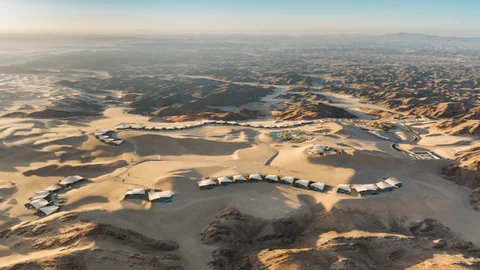 Desert surroundings with thoughtfully placed buildings and tents among sand dunes and rocky hills, under a clear sky. Designed for wellness and environmental care.