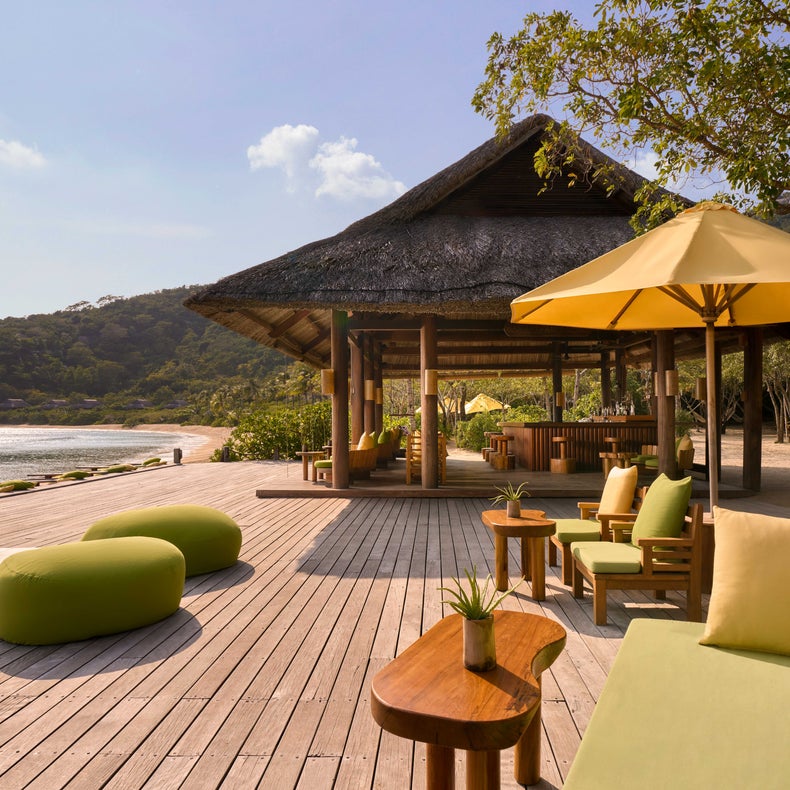 Terrasse avec des sièges verts et des parasols jaunes à côté d'un pavillon au toit de chaume, située près du rivage avec des arbres et des collines douces tout autour.