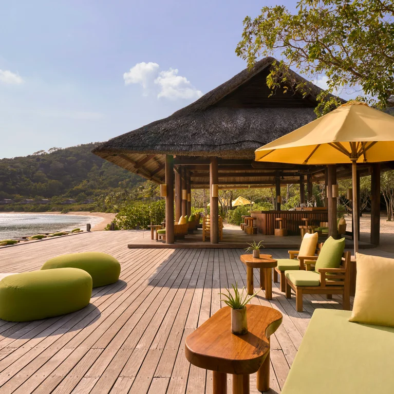 Trên ban công có những chiếc ghế bọc nệm màu xanh lá cây và những chiếc ô che nắng màu vàng, bên cạnh là một ngôi chòi lợp lá cọ, nằm sát bờ biển, xung quanh được bao phủ bởi những hàng cây xanh mát, và địa hình đồi núi thoai thoải.