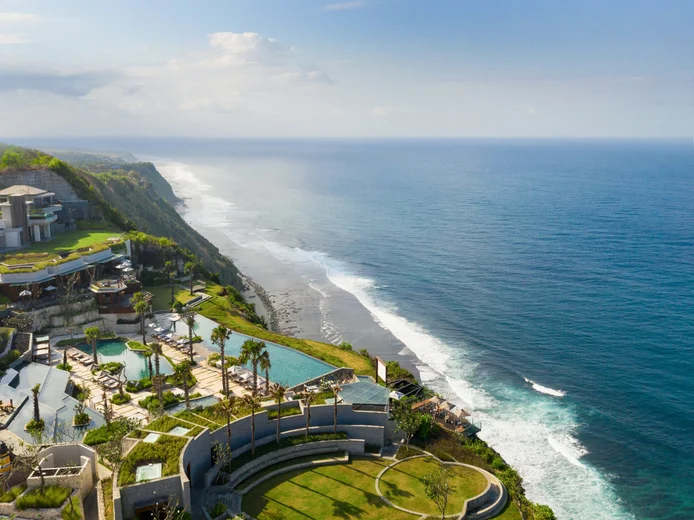Resort grounds with pools and native gardens positioned on a green cliff, offering views of the ocean and coastline under a clear sky.
