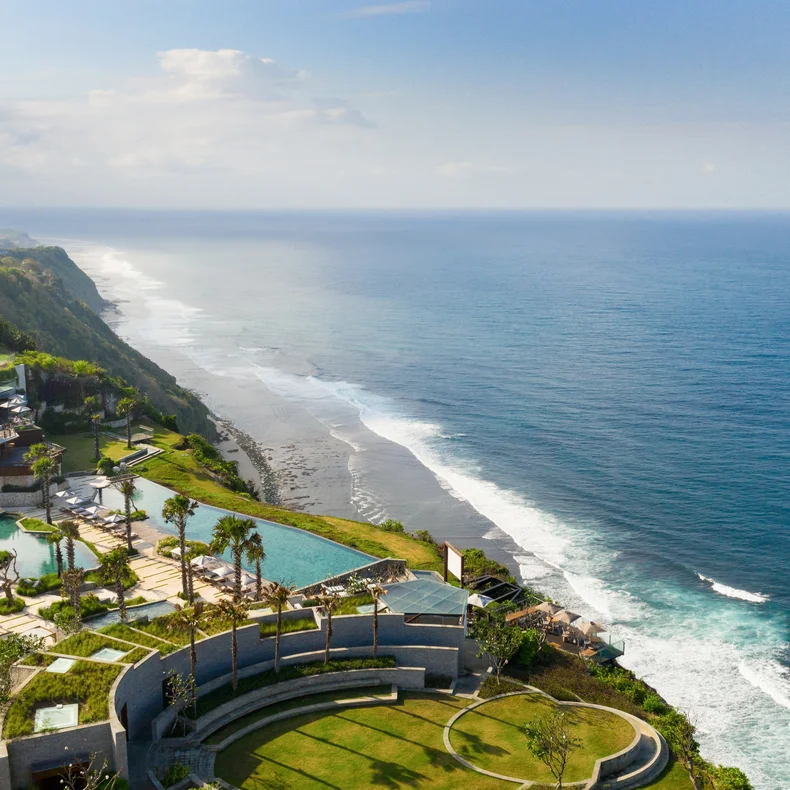 Resort grounds with pools and native gardens positioned on a green cliff, offering views of the ocean and coastline under a clear sky.