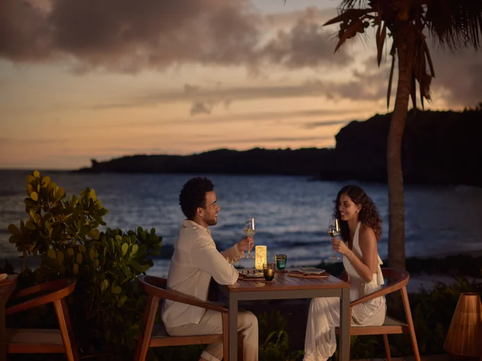 Two guests dine at an oceanfront table with beverages, surrounded by tropical greenery and gentle lighting, steps from the shore.