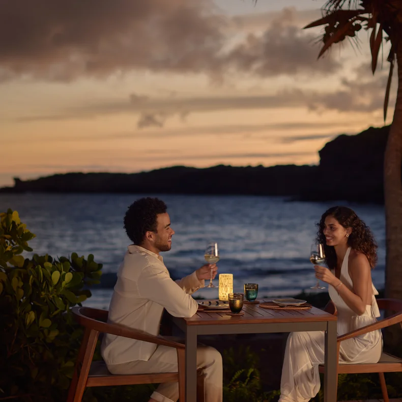 Two guests dine at an oceanfront table with beverages, surrounded by tropical greenery and gentle lighting, steps from the shore.