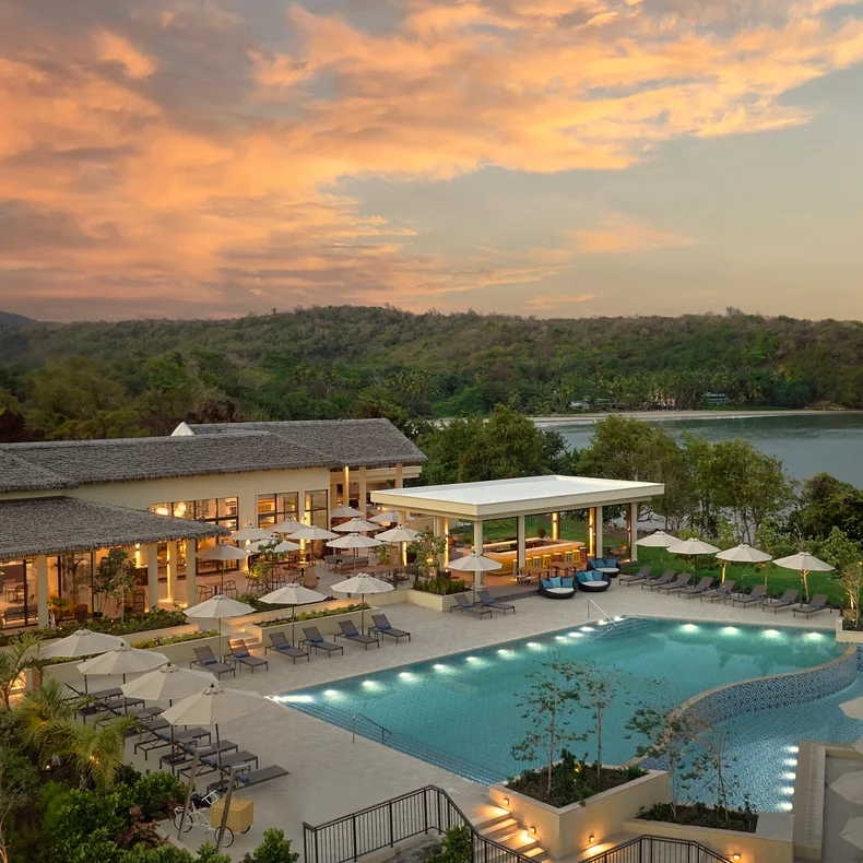 Resort pool with shaded lounge chairs faces a tranquil river and tree-covered hills at sunset, sky glowing orange above the landscape. Accessible pathways visible around pool area.