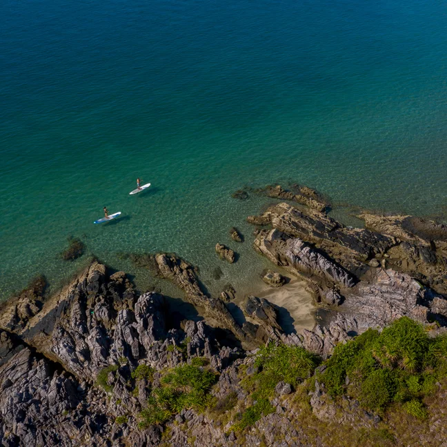 Two people paddleboard in clear blue-green water beside a rocky shoreline and lush greenery, highlighting harmony with nature and wellness activities.