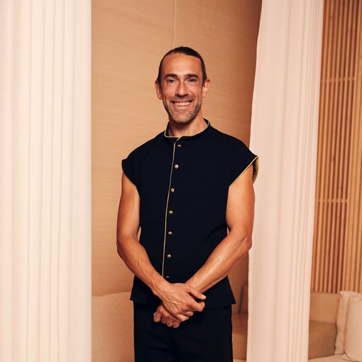 A man with tied-back hair stands indoors, wearing a black sleeveless shirt with gold buttons, positioned between white curtains in a bright, modern room.