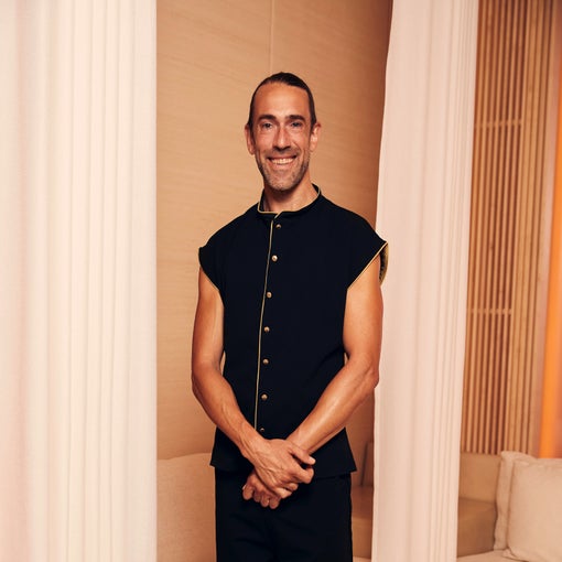 A man with tied-back hair stands indoors, wearing a black sleeveless shirt with gold buttons, positioned between white curtains in a bright, modern room.