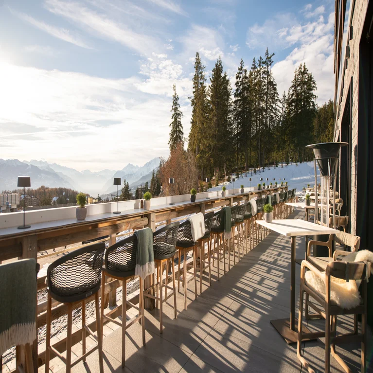 Patio with wooden tables and chairs offers views of snowy mountains and pine trees; select tables provide blankets for added comfort.