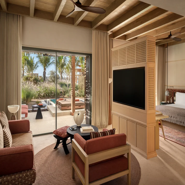 Serene guest room features natural wood ceiling, seating area, TV divider, bed, and sliding doors to a patio with pool and tropical views of palm trees.