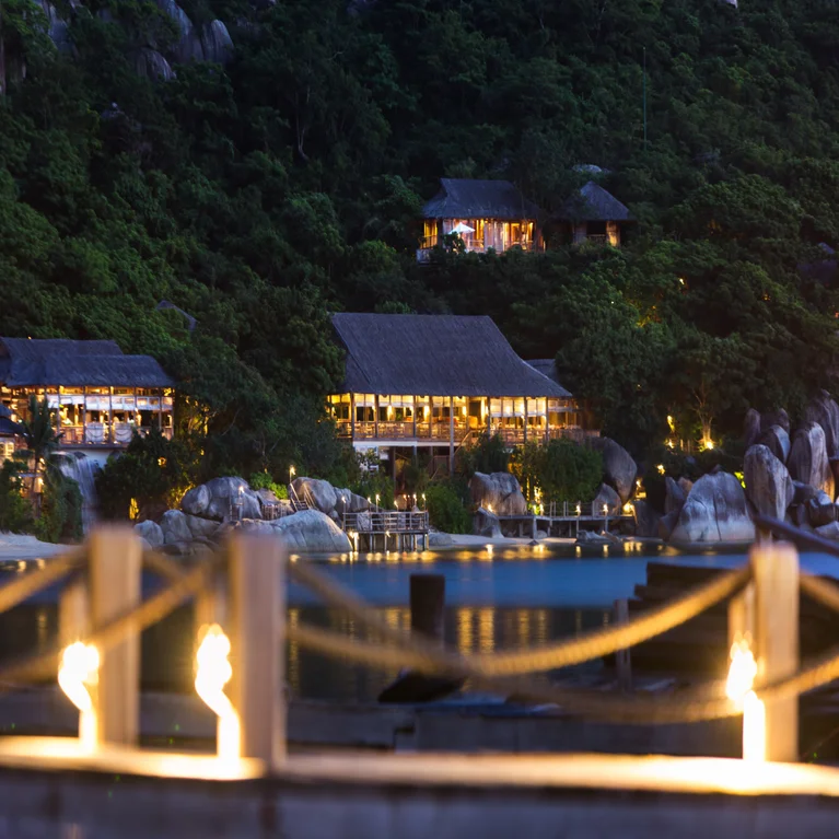 Cottages with traditional roofs amid vibrant greenery, softly lit at dusk, face a calm lake. Image viewed from behind a simple rope fence for accessibility.