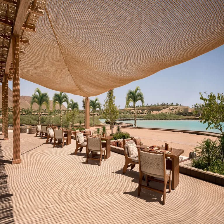 Dining area with wooden tables and chairs set beneath a mesh canopy, facing calm water and palm trees under bright natural light.