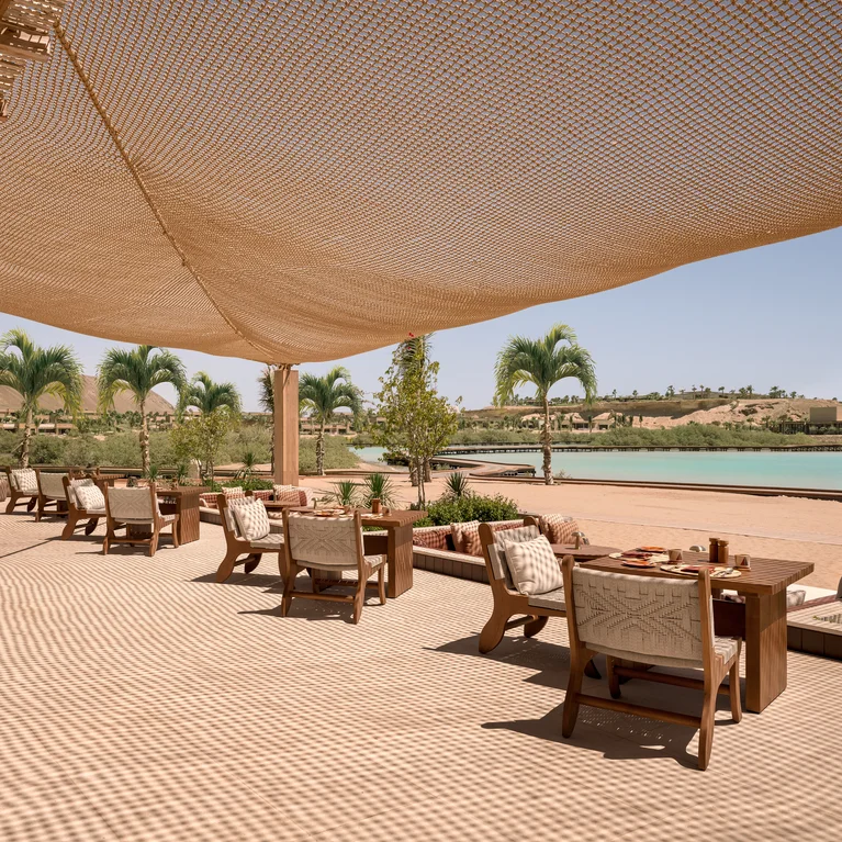 Dining area with wooden tables and chairs set beneath a mesh canopy, facing calm water and palm trees under bright natural light.