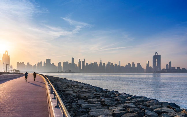An individual enjoys a morning jog on a coastal path, with rocky shoreline and contemporary city skyline visible through gentle morning mist.