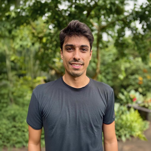 A guest with short dark hair in a black t-shirt stands outdoors, smiling softly, surrounded by vibrant greenery and lush trees.