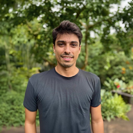 A guest with short dark hair in a black t-shirt stands outdoors, smiling softly, surrounded by vibrant greenery and lush trees.
