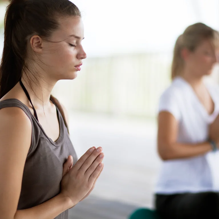 Guests practice mindful meditation together indoors, hands at heart center and eyes closed, highlighting a sense of calm and focus in an inviting wellness space.
