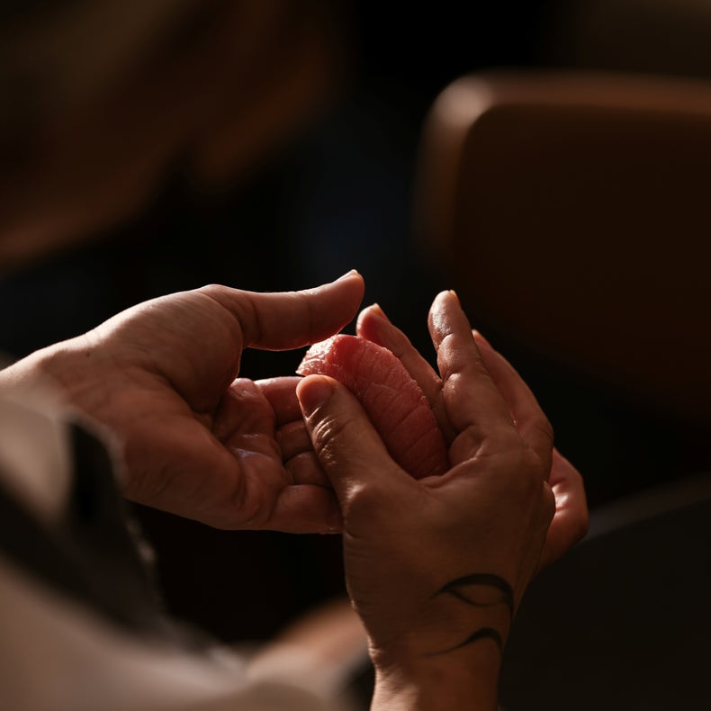 Hands gently present a piece of sushi, with a subtle tattoo on one wrist. The background is softly blurred, highlighting the focus on mindful dining.