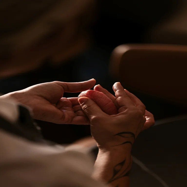 Hands gently present a piece of sushi, with a subtle tattoo on one wrist. The background is softly blurred, highlighting the focus on mindful dining.