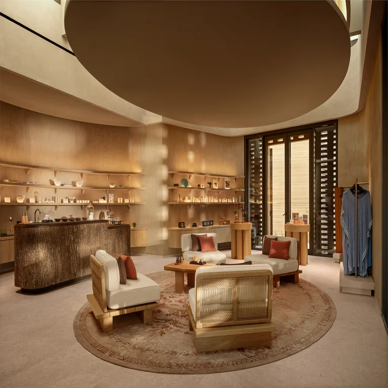 Lounge featuring cream sofas, wooden tables, decor shelves, counter, and robe on rack. Soft lighting and rounded rug support comfort and balance in a thoughtfully arranged setting.