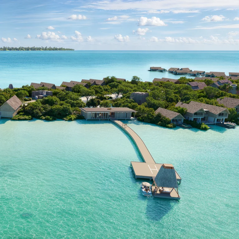 Pier over turquoise water leads to thatched-roof villas encircled by greenery, designed for relaxation and wellness beneath a clear blue sky.