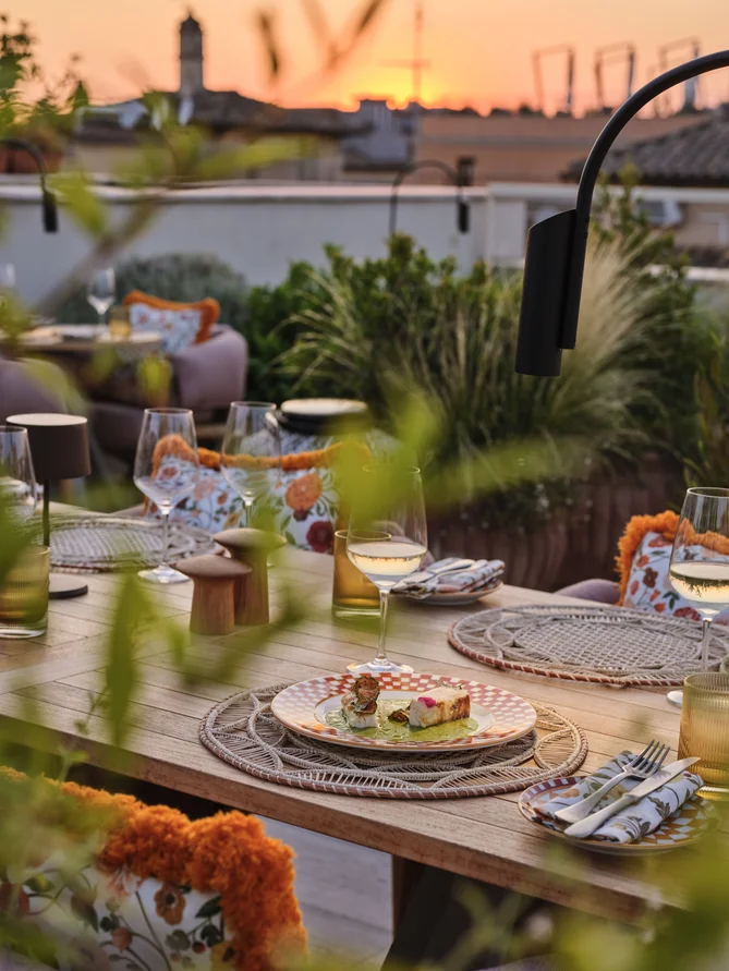 Outdoor dining table with white wine, fresh food, and floral cushions at sunset, surrounded by greenery for a serene and inviting atmosphere.