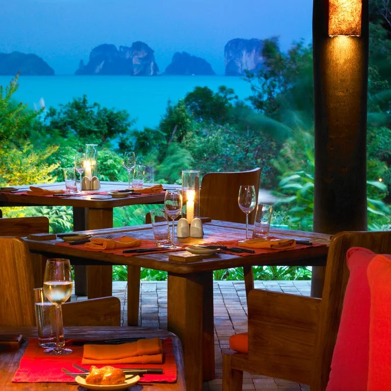 L'espace de restauration en plein air comprend des tables en bois disposées pour le dîner, éclairées par des bougies, avec vue sur la mer et les falaises au crépuscule, bordées d'une abondante verdure.