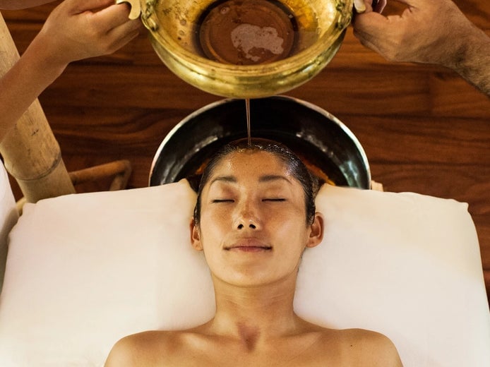 El huésped disfruta de un suave flujo de aceite en la frente durante un tratamiento de bienestar holístico, que hace hincapié en la relajación y la técnica tradicional en un tranquilo entorno de spa.