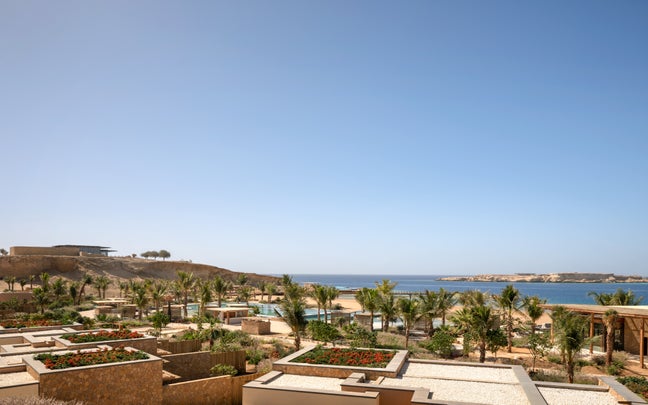 Palm trees, native gardens, and thoughtfully designed stone buildings face the blue sea, with an island and sandy cliff visible across the water beneath a clear sky.