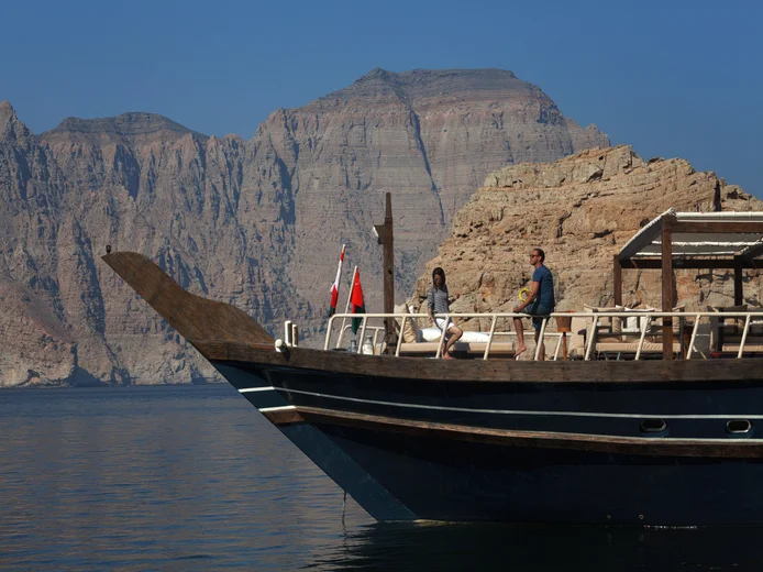 Two people travel by boat past rocky cliffs on calm water, with distant mountains and a clear sky, highlighting nature’s serenity and balance.