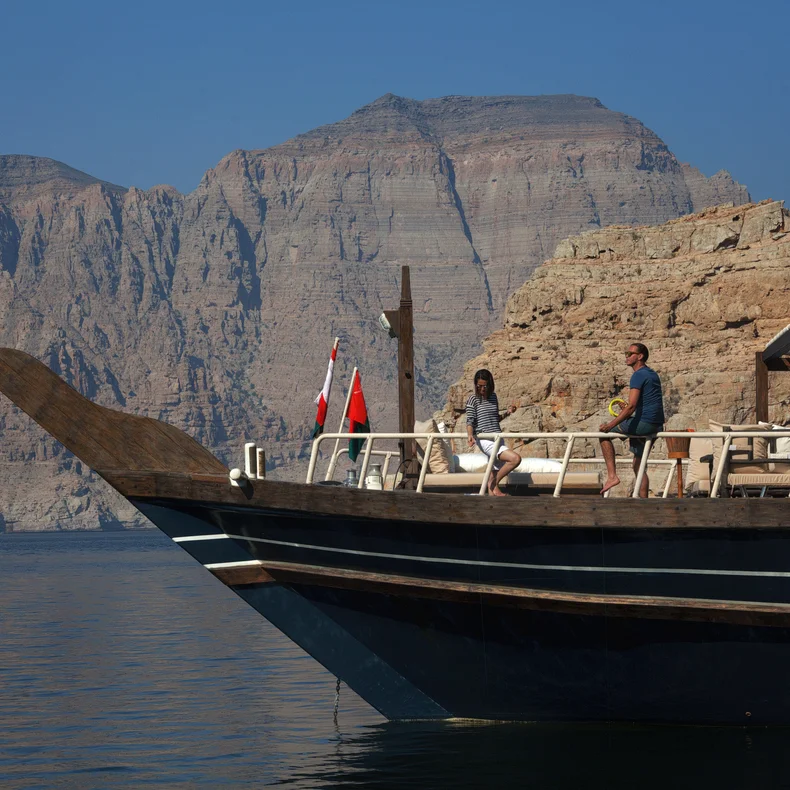 Two people travel by boat past rocky cliffs on calm water, with distant mountains and a clear sky, highlighting nature’s serenity and balance.