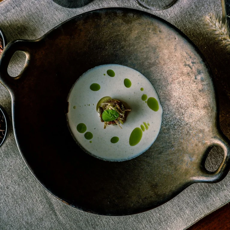 在一块中性的布上放着一碗奶油汤,中间滴着绿色的油和一个装饰品,旁边是两个装着干配料的小碗。.