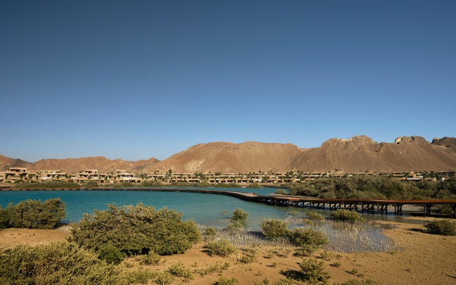 A boardwalk extends over calm, turquoise water, framed by lush green shrubs and distant mountains beneath a clear blue sky.