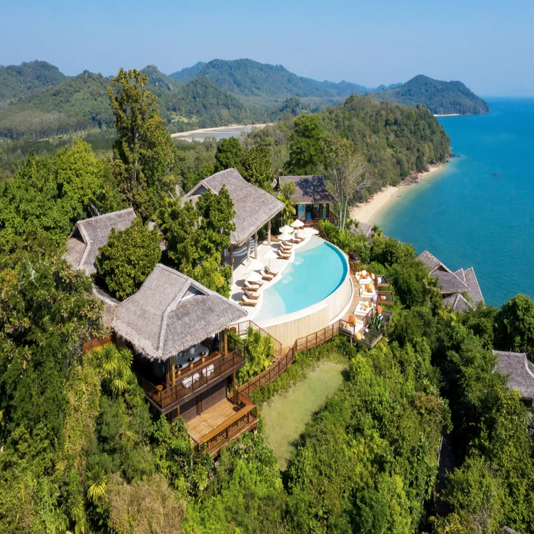 Thatched-roof villas and an infinity pool set beside green forest, sandy beach, and turquoise sea beneath a clear blue sky. Designed for wellbeing and harmony with nature.