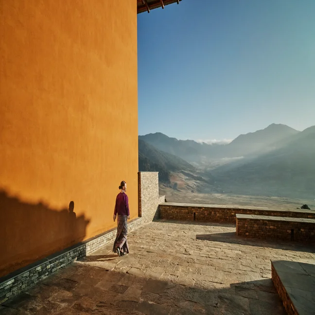 A guest strolls along a stone terrace with an orange wall, overlooking a tranquil mountain valley under a clear blue sky.