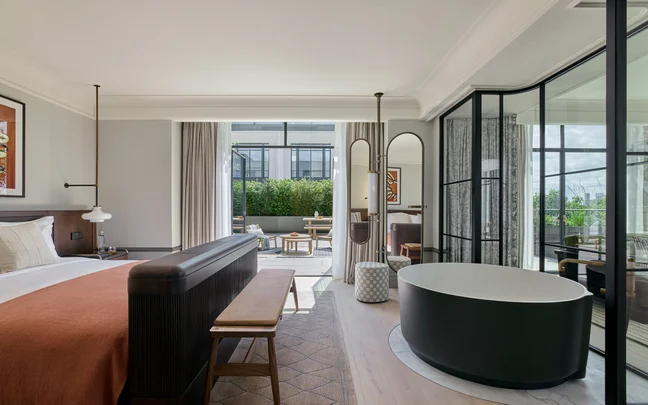 Guest room featuring a king bed, seating bench, freestanding round tub, and glass doors opening onto a terrace with outdoor chairs and views of garden greenery.