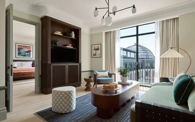 Living room with large window, green sofa, dark wood TV console, round coffee table and view of a glass-domed building. Bedroom visible through open doorway.