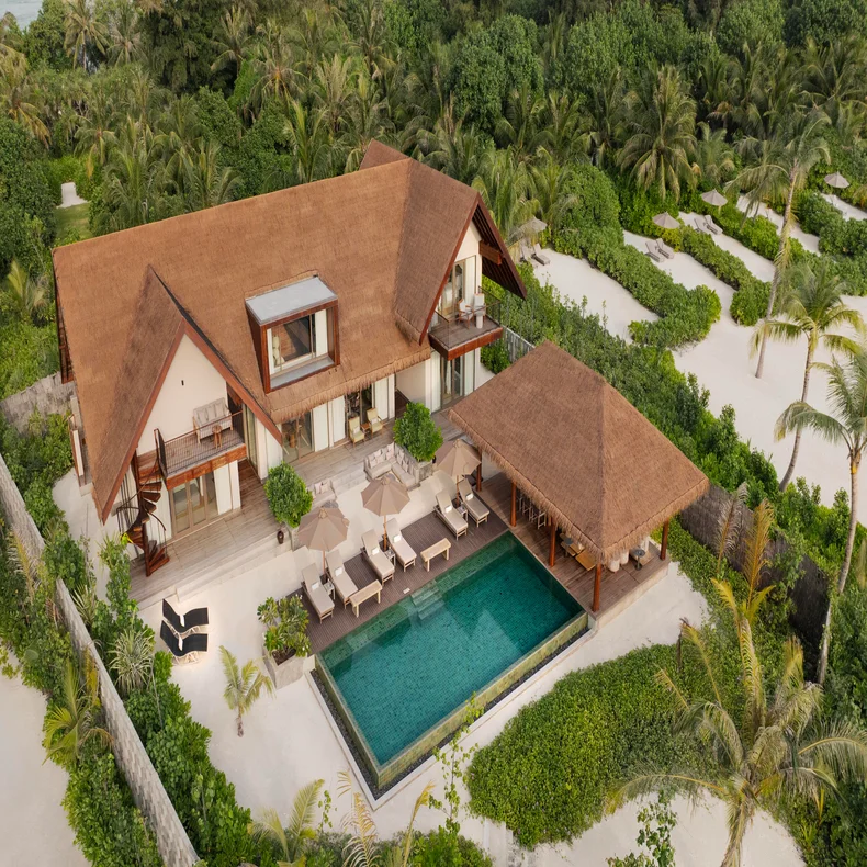 Villa with thatched roof, private pool, and sun loungers sits among abundant tropical plants and winding sandy paths. Thoughtfully designed for wellness and sustainability.