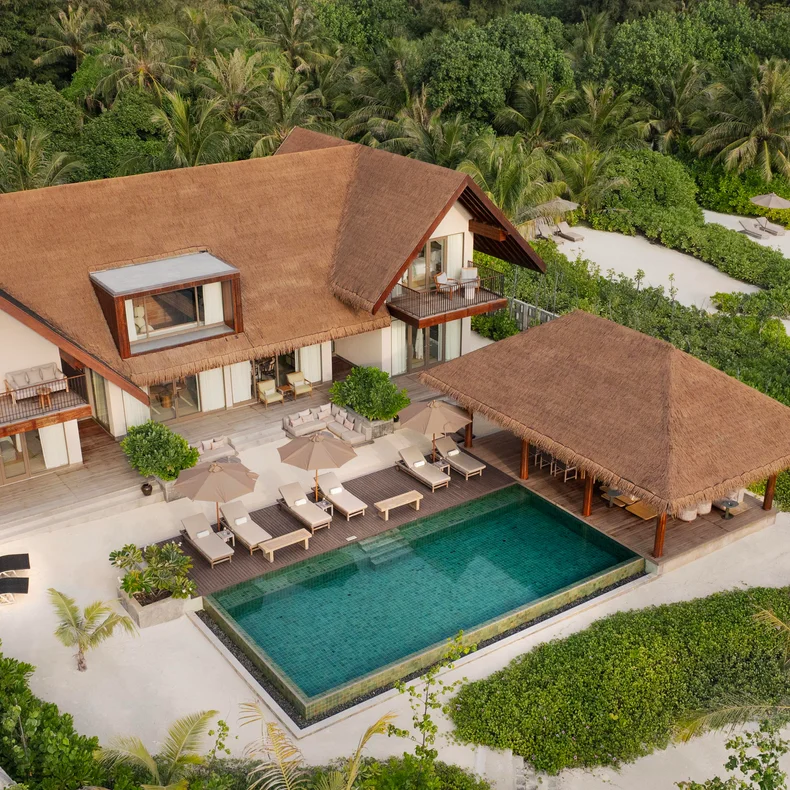 Villa with thatched roof, private pool, and sun loungers sits among abundant tropical plants and winding sandy paths. Thoughtfully designed for wellness and sustainability.