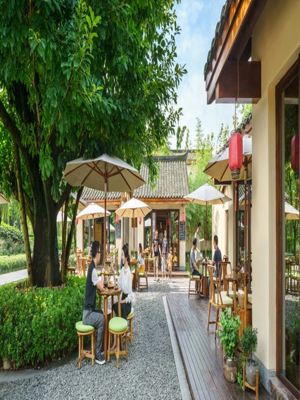 Guests enjoy shaded outdoor seating in a tranquil garden café with wooden chairs, surrounded by trees and lanterns on a bright day.