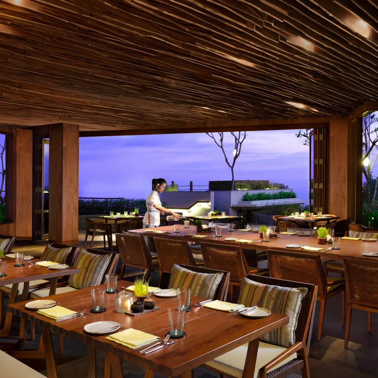 مساحة مفتوحة لتناول الطعام في الهواء الطلق تتميز بلمسات خشبية طبيعية وطاولات مرتبة بعناية ووسائد مخططة وموظفة تحضّر للضيوف بجانب نافذة واسعة مطلة على الهواء الطلق.