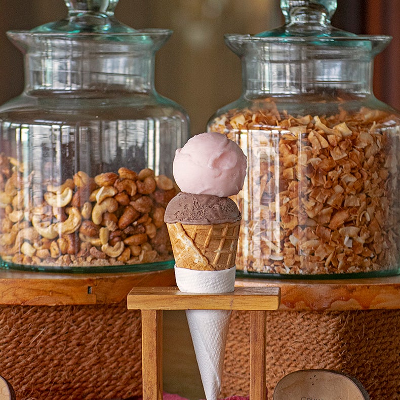 Ice cream cone with pink and brown scoops placed before glass jars of cashew nuts and toasted coconut flakes on a neutral surface.