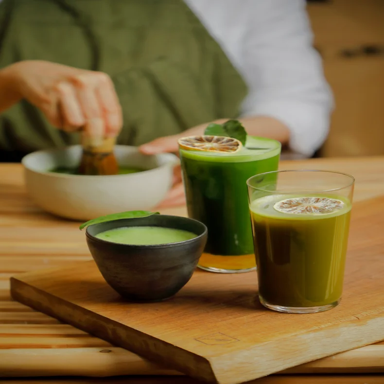 Wellness-inspired matcha beverages, served in glasses with dried lime and a black cup on wood, as matcha is freshly whisked in the background.