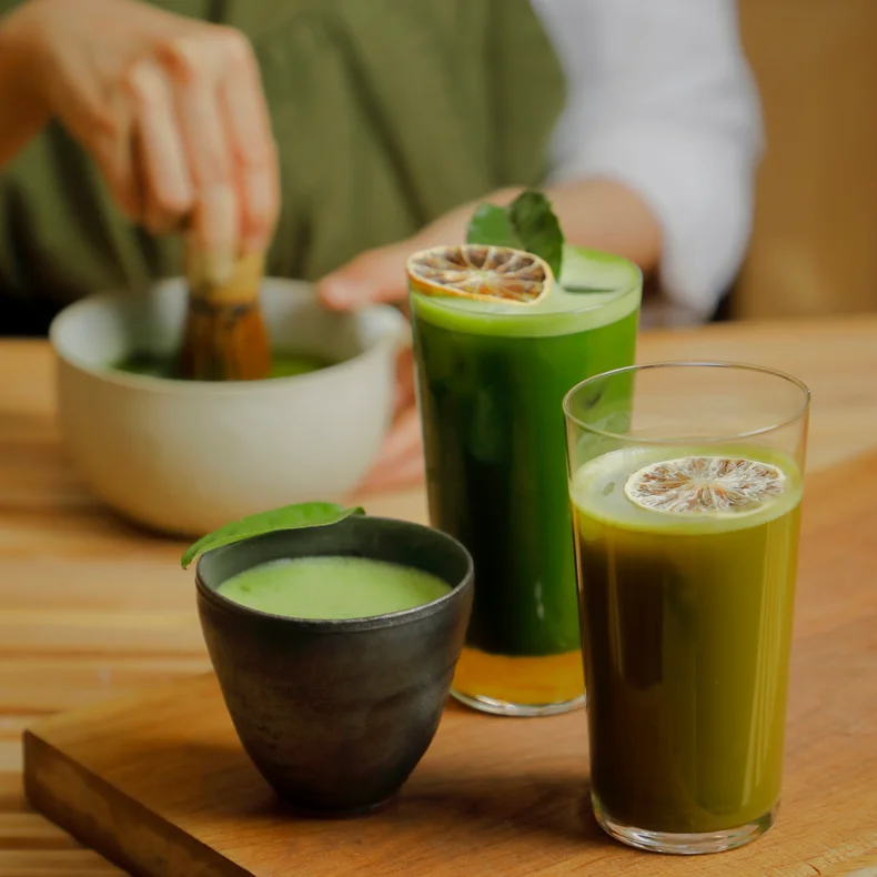 抹茶が泡立てられるのをバックに、ドライライム入りのグラスと木の上に置かれた黒いカップで提供されるウェルネスにインスパイアされた抹茶ドリンク。.