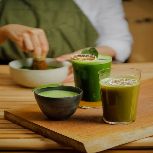 Wellness-inspired matcha beverages, served in glasses with dried lime and a black cup on wood, as matcha is freshly whisked in the background.
