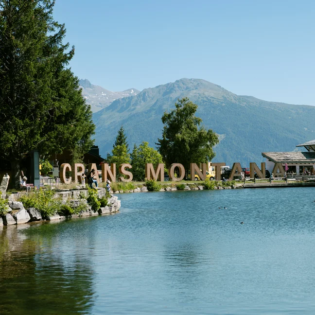 Lac avec un panneau « CRANS MONTANA », bordé d'arbres et de montagnes. Des bâtiments modernes sont visibles à droite. Le ciel bleu dégagé met en valeur les éléments naturels et architecturaux.
