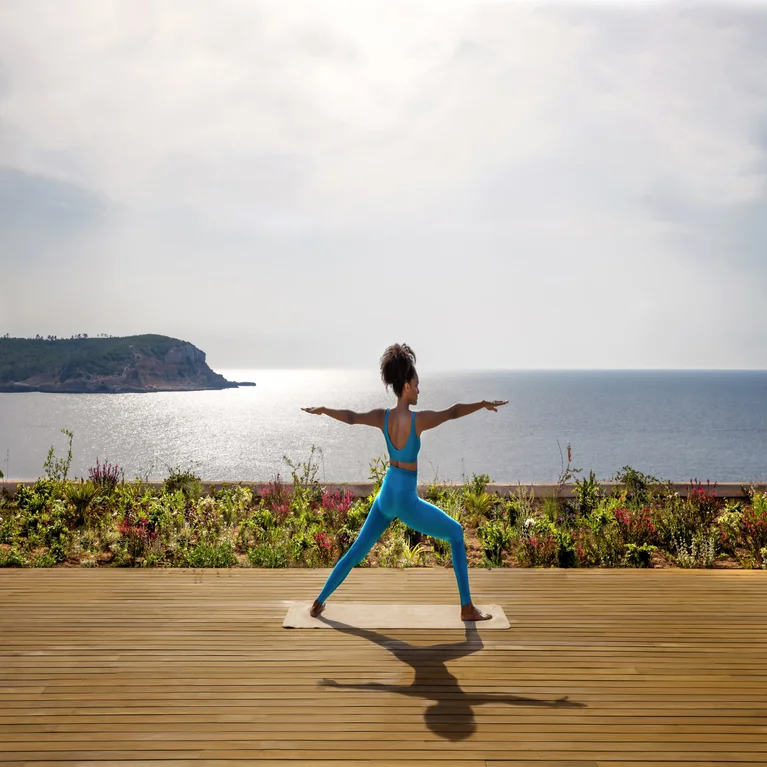 Individual in a blue outfit holds an open-arm yoga pose on a wooden deck above the ocean, with sunlight reflecting on the water in the background.
