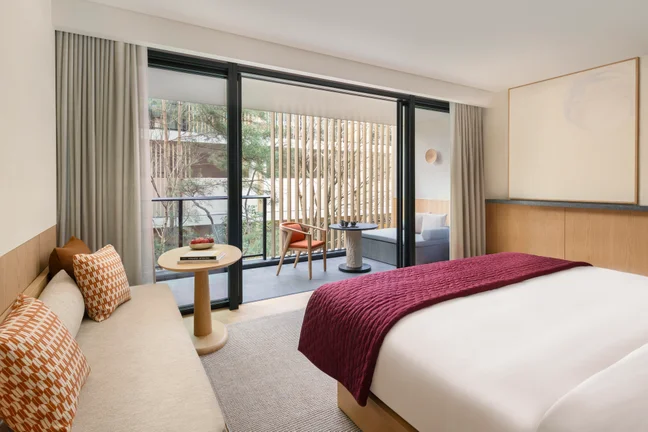 Bright guest room with king bed, sofa, low table, and private balcony set with two chairs. Large glass doors open to natural light; calming neutral palette accented by a burgundy throw.