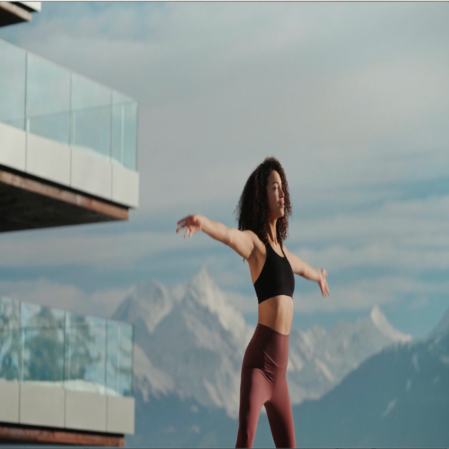 Guest practices yoga on a balcony featuring glass railing, with mountain views and blue skies, reflecting Six Senses’ commitment to wellness and connection with nature.