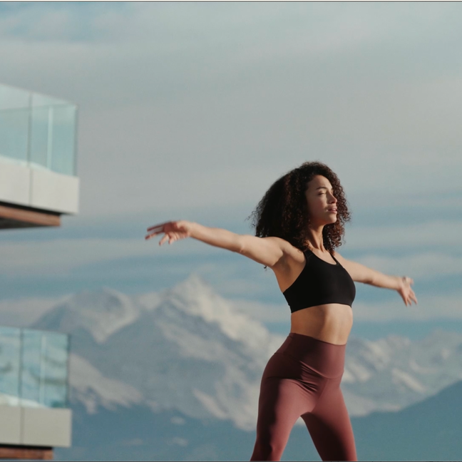 Guest practices yoga on a balcony featuring glass railing, with mountain views and blue skies, reflecting Six Senses’ commitment to wellness and connection with nature.