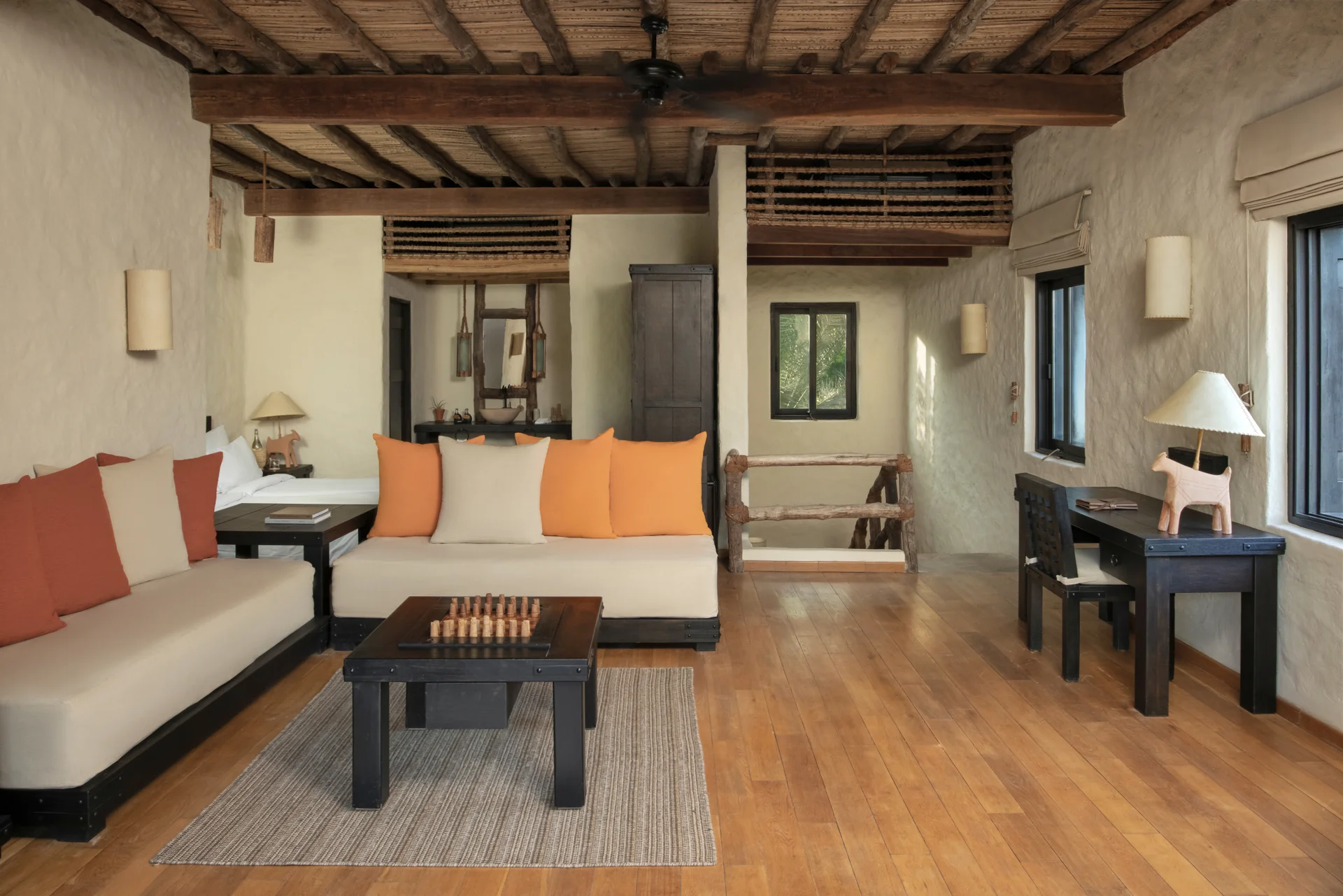 The upper living floor of a two-bedroom duplex villa, built-in banquettes cushioned in cream with orange and rust bolsters, a low carved games table and a dark writing desk beneath heavy exposed beams.
